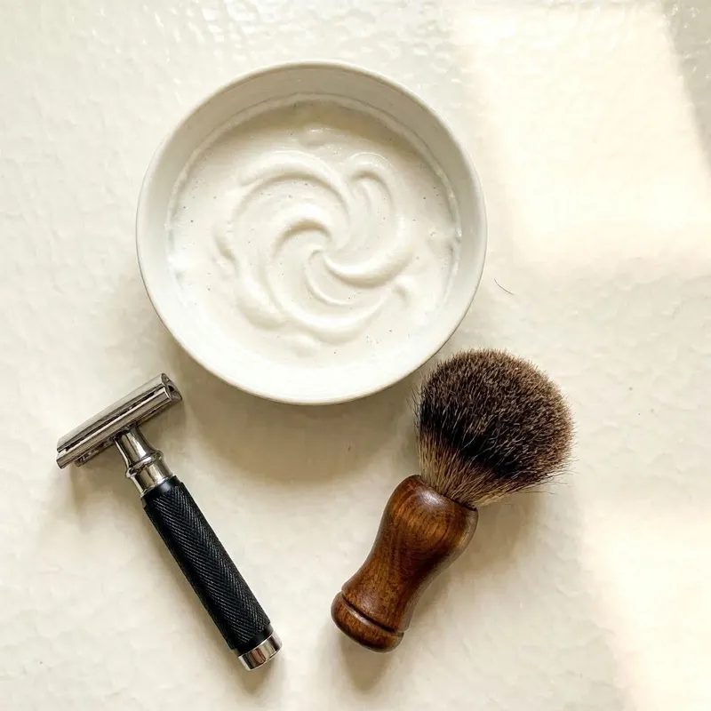 shaving essentials for coarse or curly hair including a safety razor, a shaving brush, and a bowl of rich lather, with subtle curved foam patterns on a clean white bathroom surface